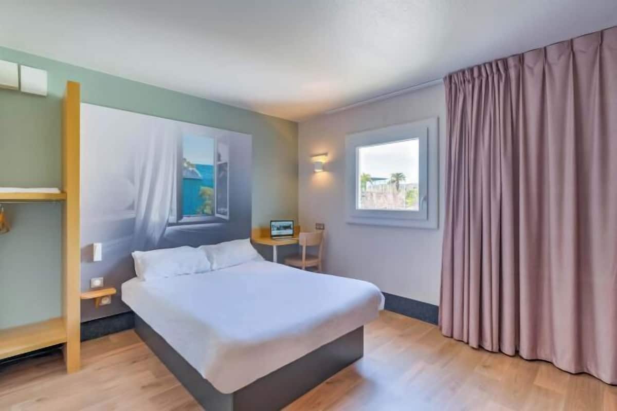 A hotel room with a bed, a desk, a chair, a window with curtains, and a painting on the wall.