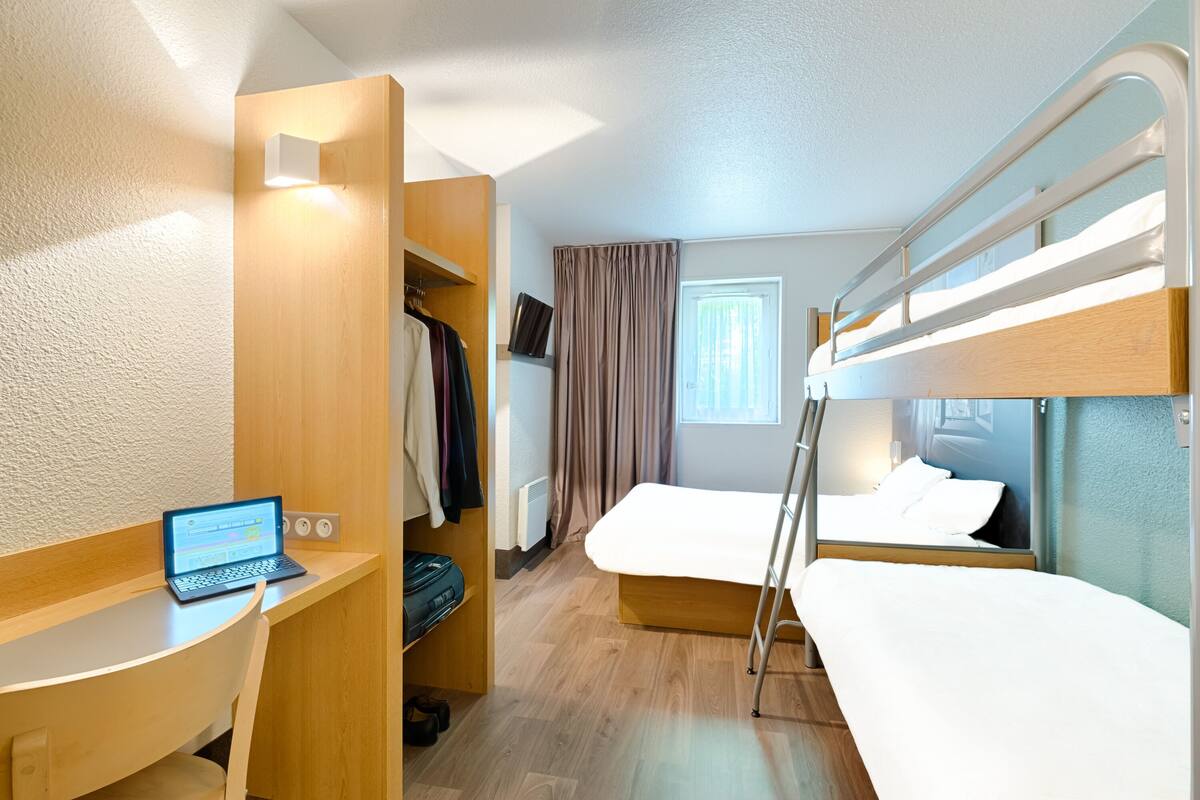 A compact hotel room with a bunk bed, a desk with a laptop, a wardrobe, and a window with curtains.