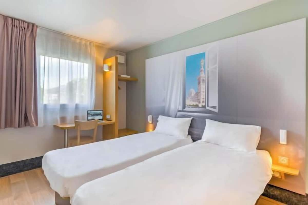 A hotel room with two beds, a desk, a chair, and a painting on the wall.