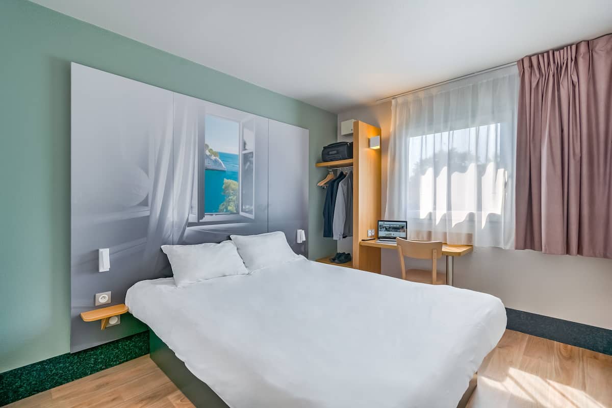 A modern hotel room with a large bed, a desk with a laptop, a chair, and a view of the sea through a window.