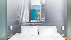 A bedroom with a bed, white pillows, a nightstand, and a window with a view of a coastal landscape. - B&B HOTEL Marseille La Valentine (Marseille)