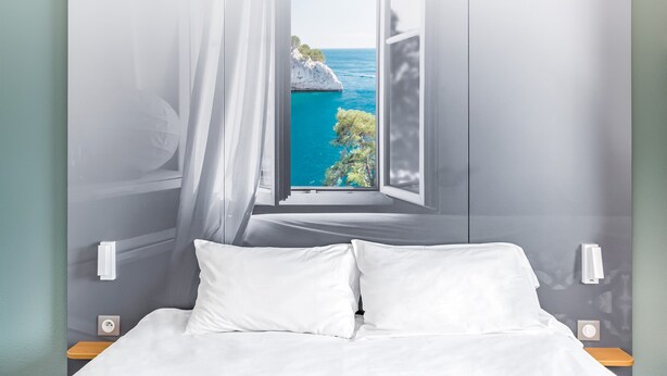 A bedroom with a bed, white pillows, a nightstand, and a window with a view of a coastal landscape. - B&B HOTEL Marseille La Valentine (Marseille)