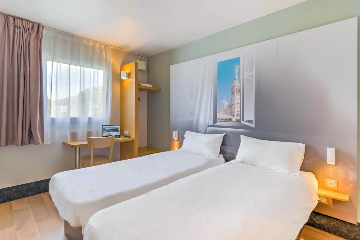 A hotel room with two beds, a desk, a chair, a computer, and a painting on the wall.