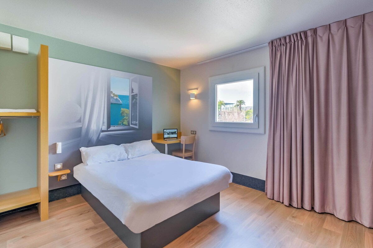 A hotel room with a bed, a desk, a chair, a window with curtains, and a wall-mounted light.