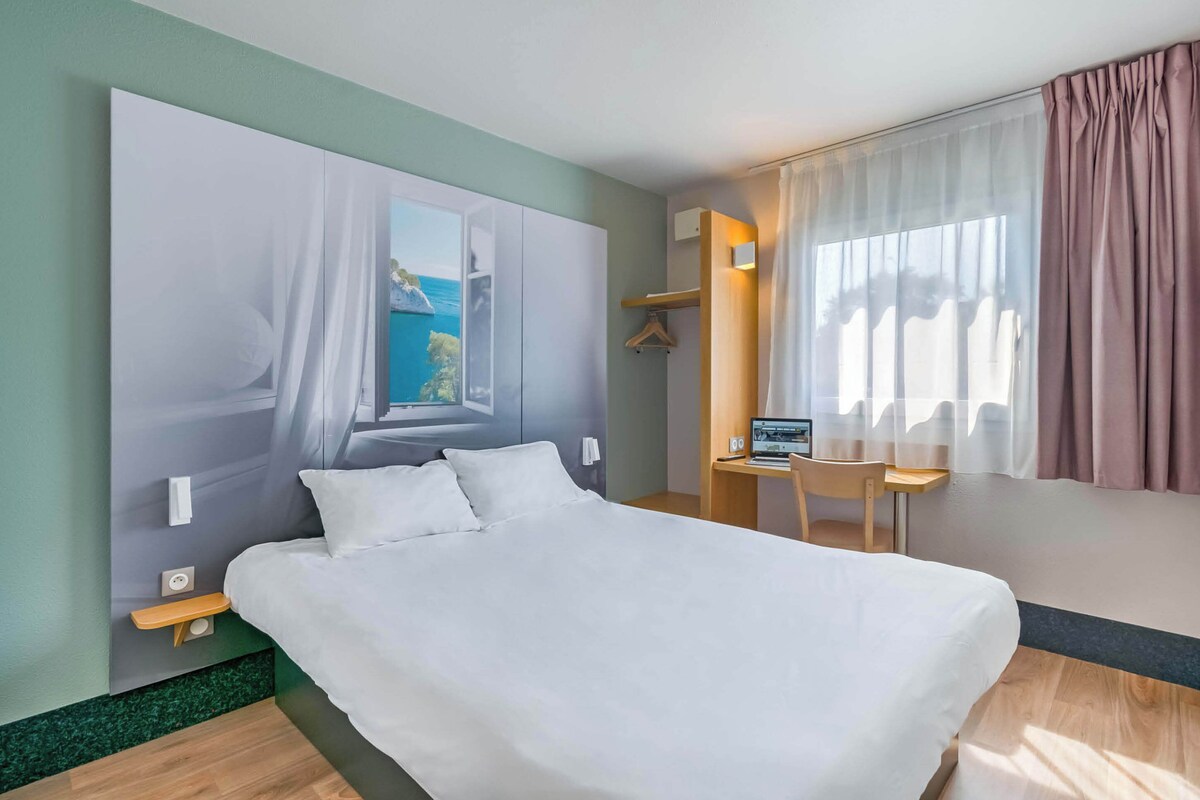 A hotel room with a large bed, a desk, a chair, a window with curtains, and a painting on the wall.