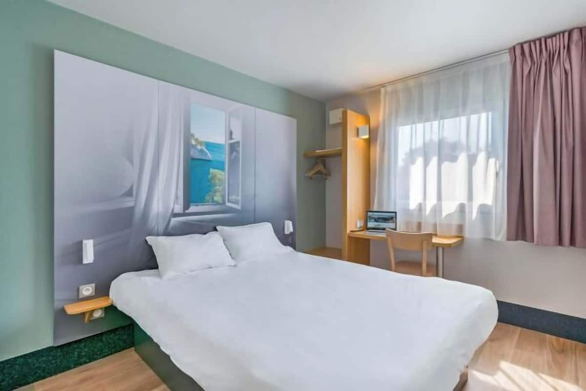 A hotel room with a bed, a desk, a chair, a window with curtains, and a view of the outside.