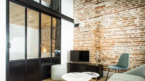 Un salon moderne avec un mur en briques, un meuble de rangement noir pour appareils électroniques, une table ronde blanche et un canapé vert.