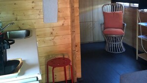 Un intérieur en bois comprenant un coin cuisine, un tabouret rouge, une chaise en osier avec un coussin et une armoire.