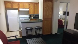 A compact kitchen with wooden cabinets, a refrigerator, stove, and a microwave. There are two stools and a small table. A bed with a red blanket is visible in the adjacent room. - Travel Inn & Suites (Atlanta)