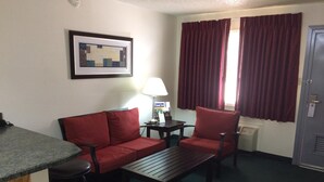 A hotel room with a red sofa, a wooden coffee table, a red armchair, a small table with a lamp, a framed picture on the wall, and a window with red curtains. - Travel Inn & Suites (Atlanta)