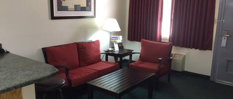 Habitación de hotel con un sofá rojo, una mesa de centro de madera, un sillón rojo, una mesita con lámpara, un cuadro enmarcado en la pared y una ventana con cortinas rojas.
