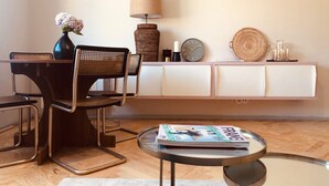 Una sala de estar moderna con una mesa de comedor, sillas, una mesa de centro, una revista, un jarrón con flores, una lámpara, un reloj y un plato decorativo sobre un mueble blanco.