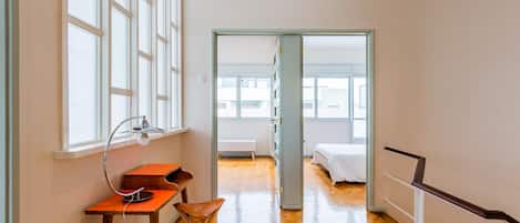 A room with a wooden floor, a desk with a lamp, a chair, and a bed.