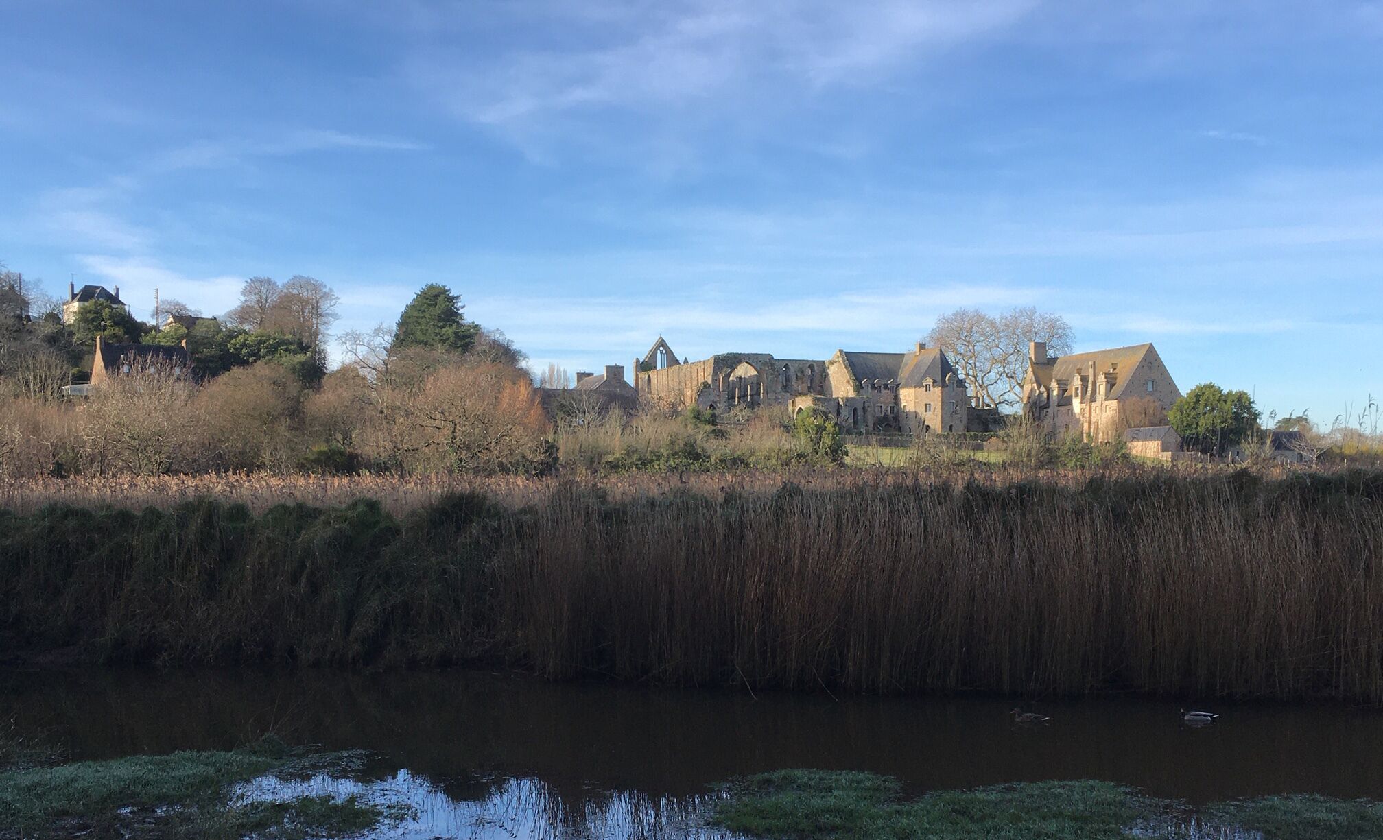 A historic building complex with multiple structures, including a large church with a pointed roof and smaller buildings with gabled roofs, situated by a river.