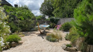 En trädgård med sand, en hängmatta, olika växter och en stenmur.