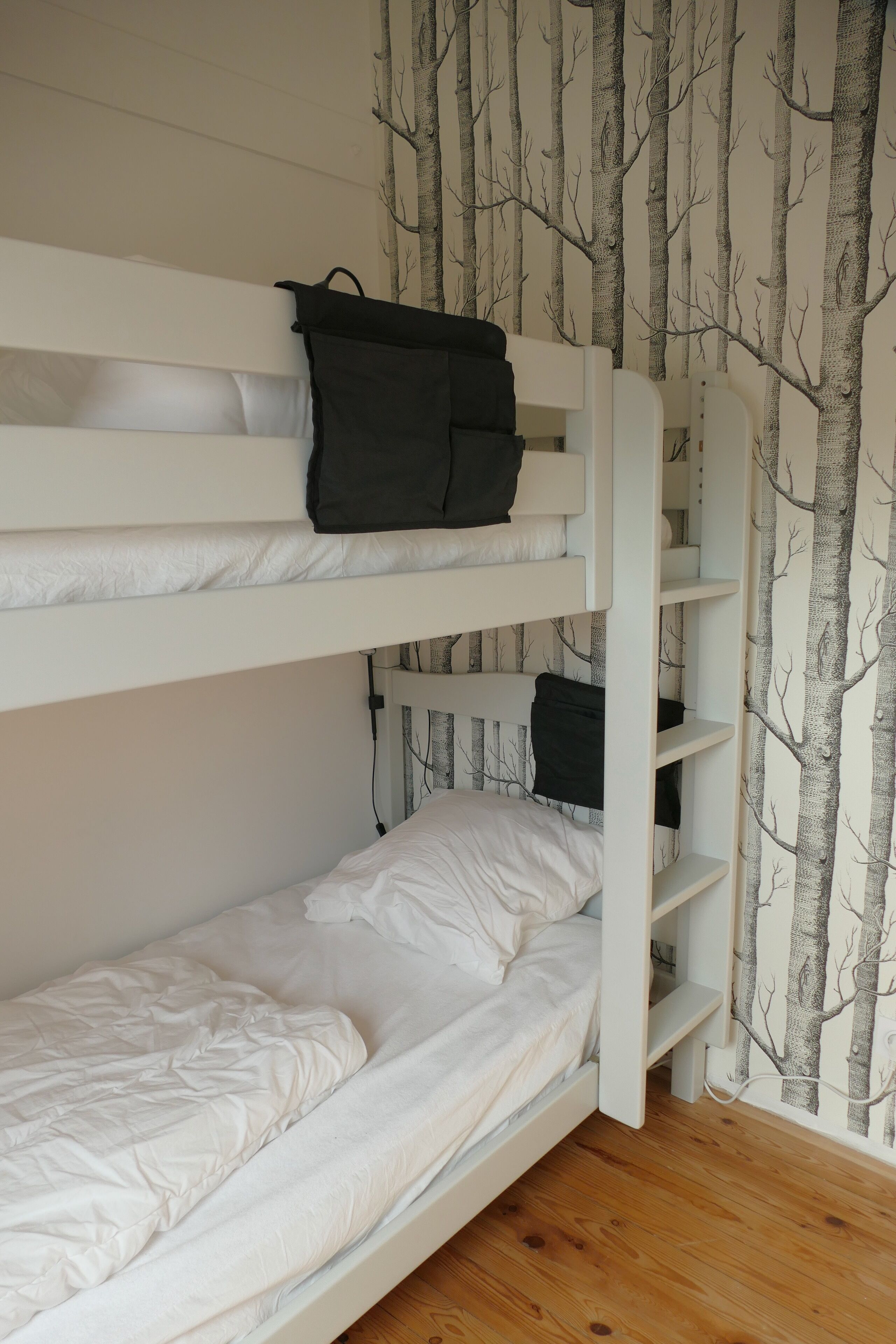 Une chambre avec un lit superposé, un papier peint à motifs d’arbres, un plancher en bois et une tête de lit blanche.