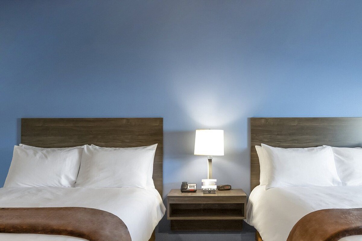 A hotel room with two beds, a nightstand with a lamp, and a small table with a phone and a card.