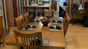 A dining room with a wooden table set for six, surrounded by wooden chairs. A chandelier hangs from the ceiling, and a kitchen is visible in the background. - Centrally located for all your activities! (Colville)