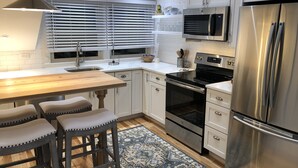 A modern kitchen with white cabinetry, a stainless steel refrigerator, a microwave, and an oven. There are bar stools and a wooden table. - Wonderful Mid Island Gem (Nantucket)
