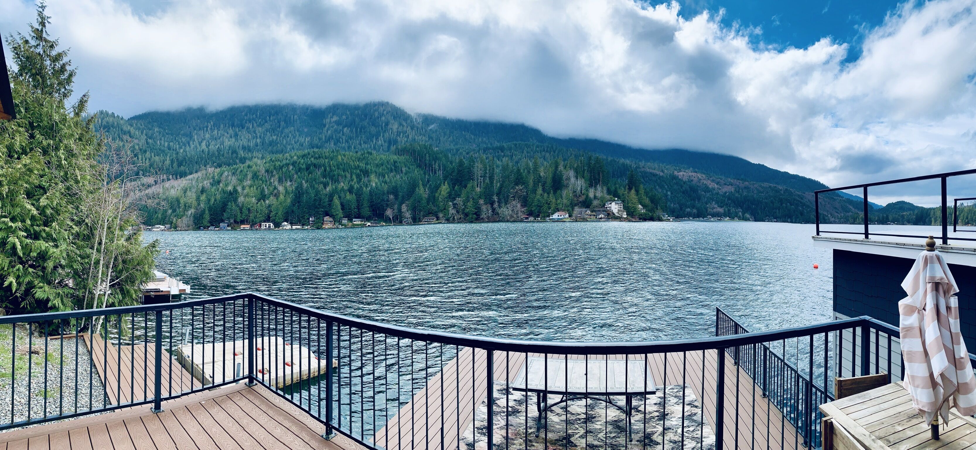 A lakeside deck with a view of a lake, mountains, and a house across the water.