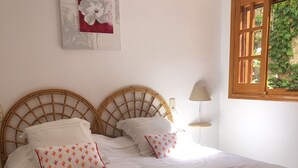Un lit double avec une couvre-lit blanche et deux oreillers, une tête de lit en bois, une table de chevet avec une lampe et une fenêtre donnant sur un paysage verdoyant.