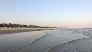 A beach with a sandy shore and waves gently lapping at the edge. In the background, there are houses along the coastline. - Short, pleasant walk to the beach! Two bedrooms, two baths. Dogs OK. (Kiawah Island)