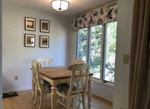 Un coin repas avec une table en bois et quatre chaises, des tableaux encadrés au mur et une fenêtre donnant sur un paysage verdoyant.