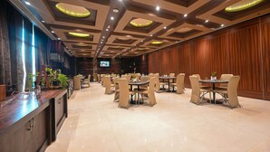 Amplia zona de comedor de hotel con varias mesas y sillas, un mostrador de recepción de madera y una pantalla de televisión en la pared.