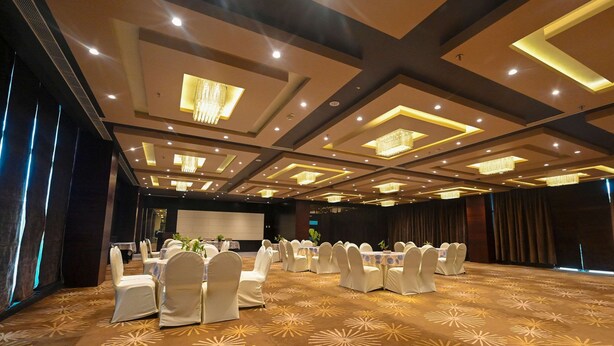 Un salón de banquetes con mesas redondas y sillas blancas, un escenario y una alfombra estampada.