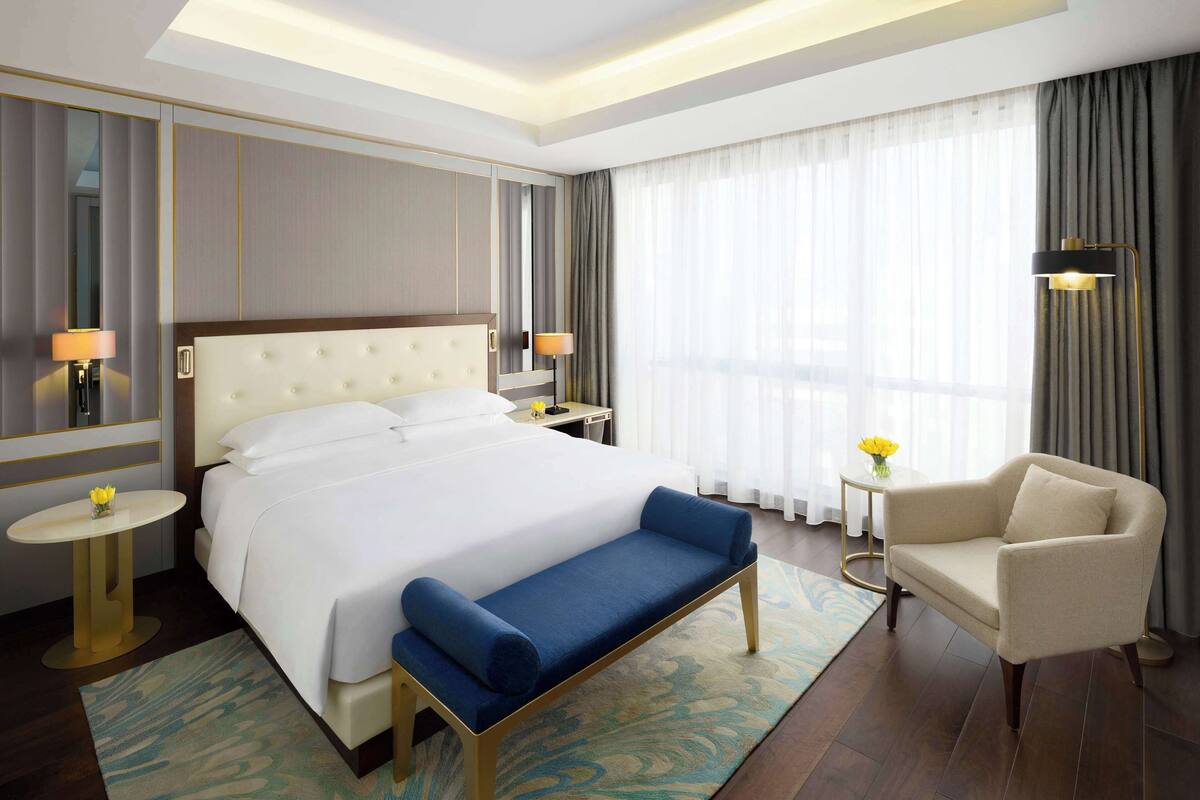 A hotel room with a large bed, a bench, a chair, a bedside table, a lamp, and a window with curtains.