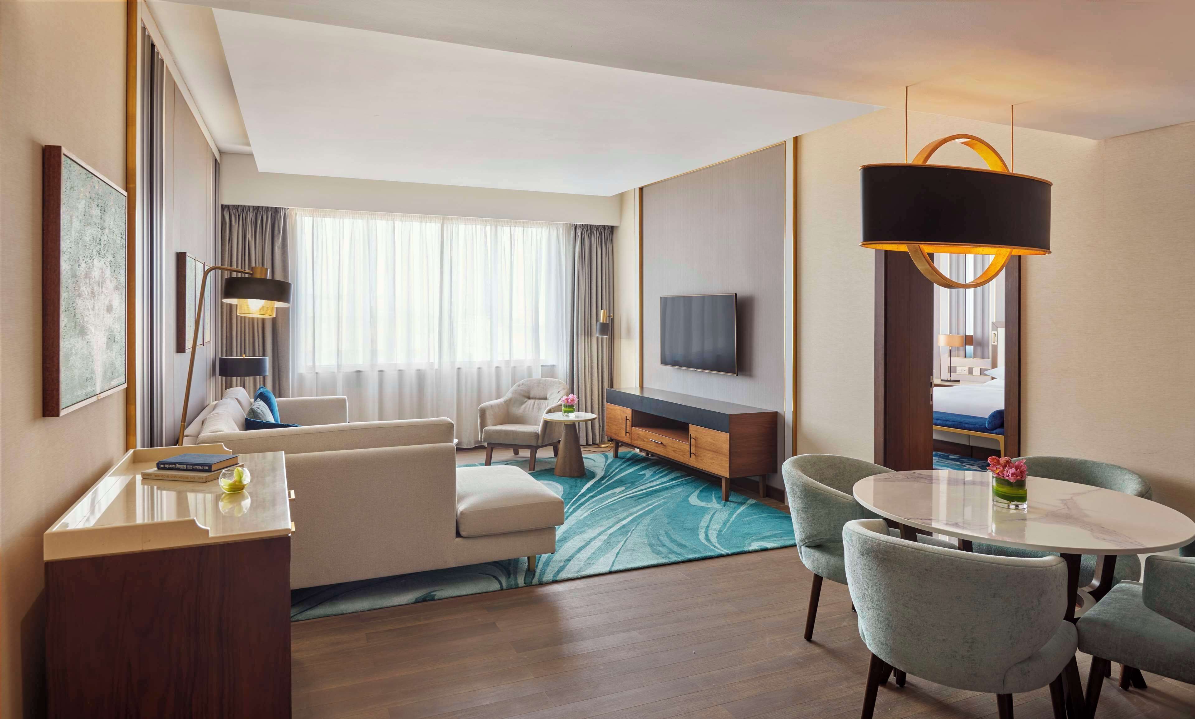 A hotel room with a sofa, armchairs, a dining table, a TV, and a large window with curtains.
