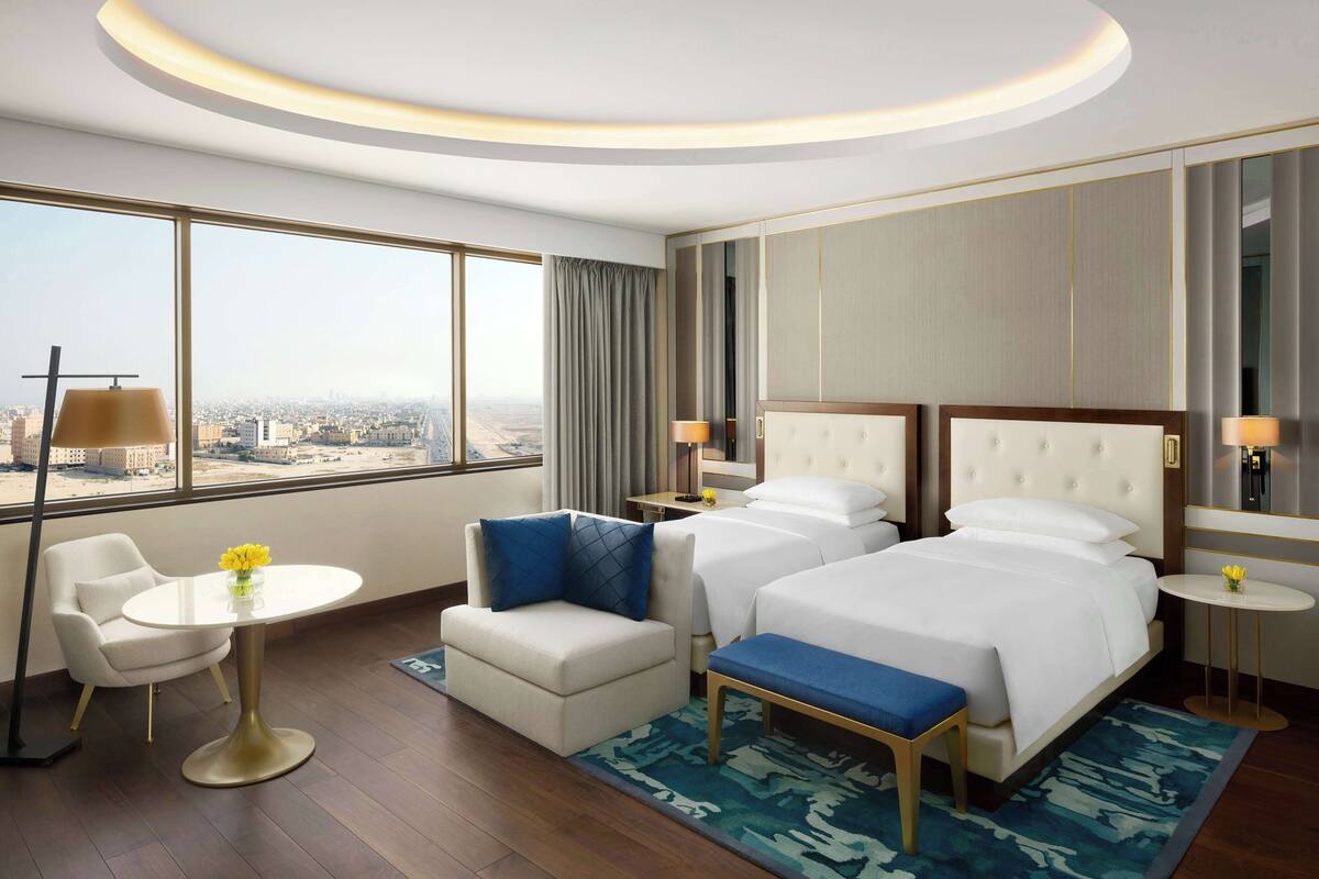 A hotel room with a large window, two beds, a sofa, a small table, and a bedside table with lamps.