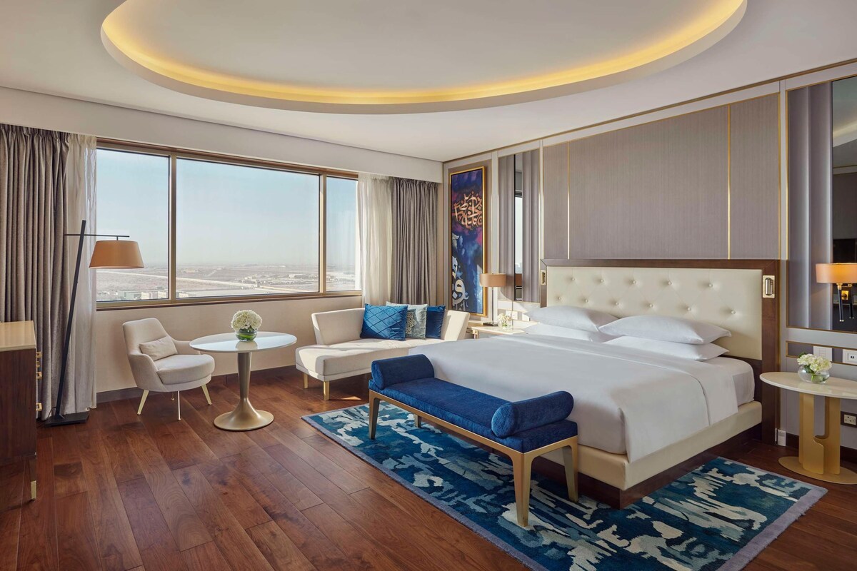 A hotel room with a large bed, a desk, a chair, a small table, and a TV.