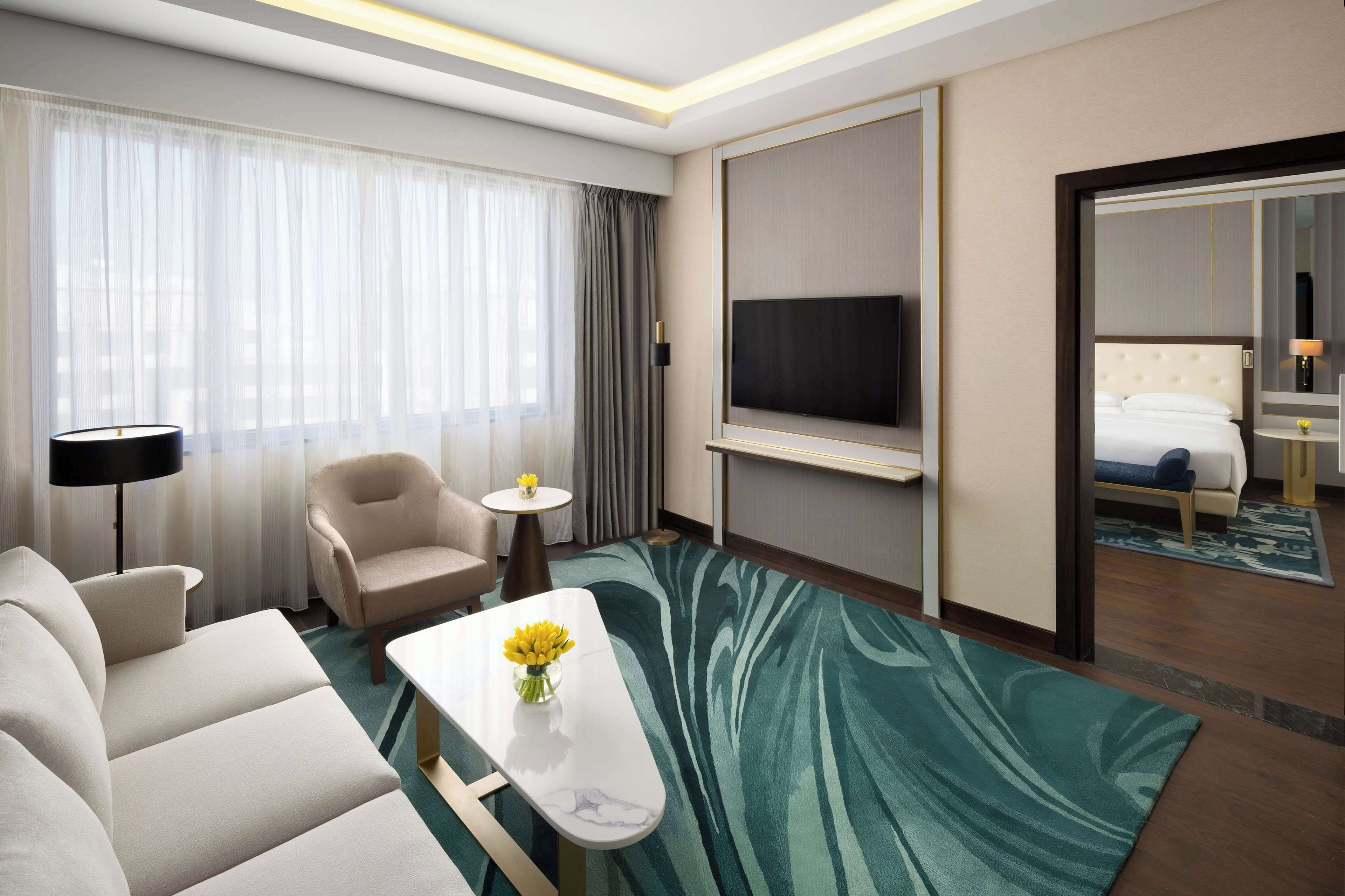 A hotel room with a sofa, armchair, small table with flowers, a TV, and a bed with a nightstand.