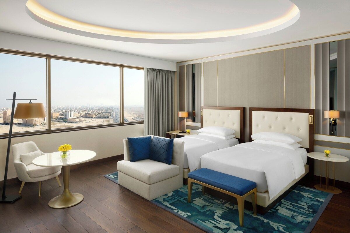 A hotel room with a large window, two beds, a small table, a chair, and a bench.