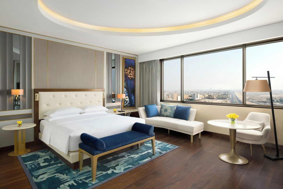 A modern hotel room with a large bed, a sofa, a small table, and a view of the city.