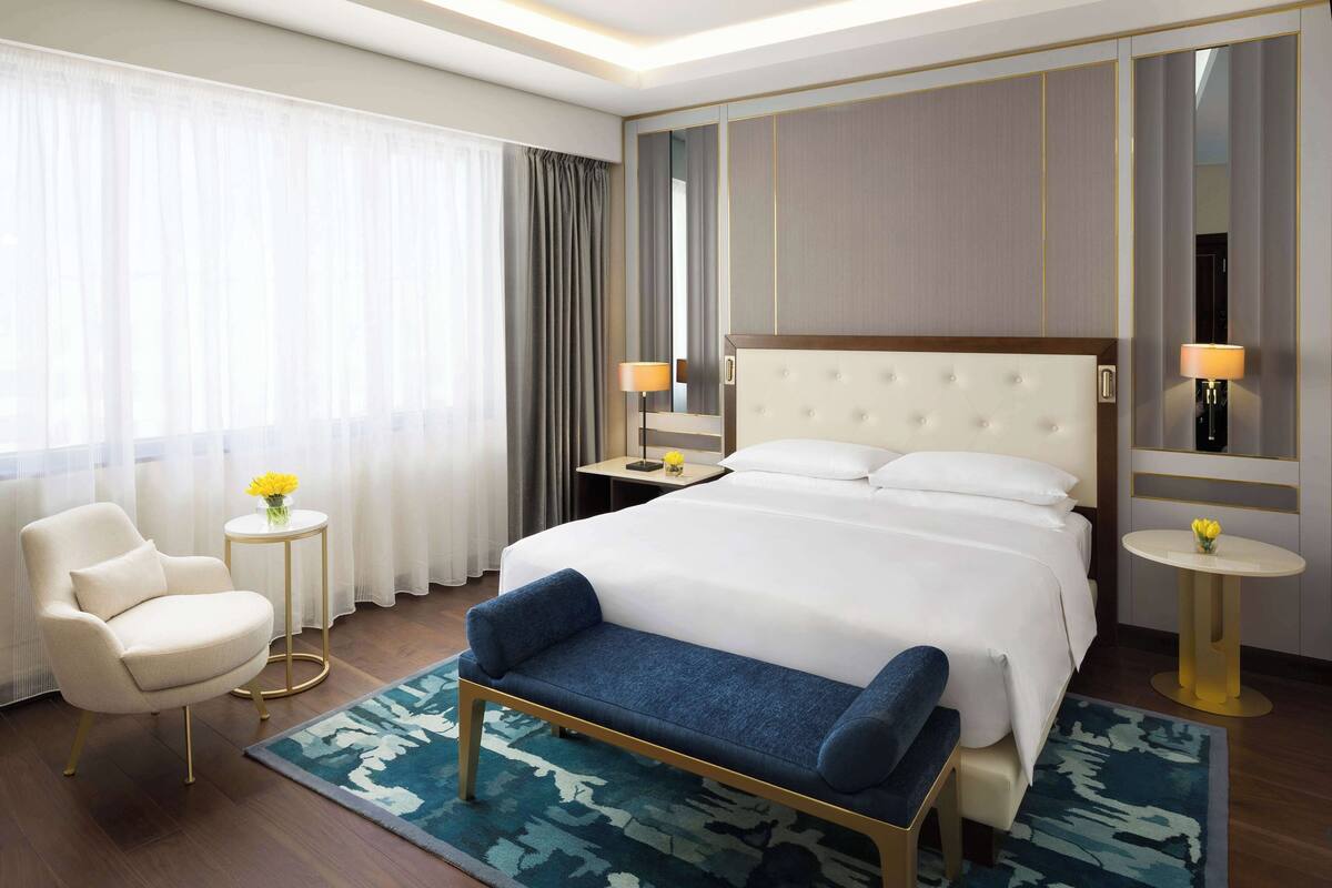 A hotel room with a large bed, a chair, a small table with a lamp, and a mirror.