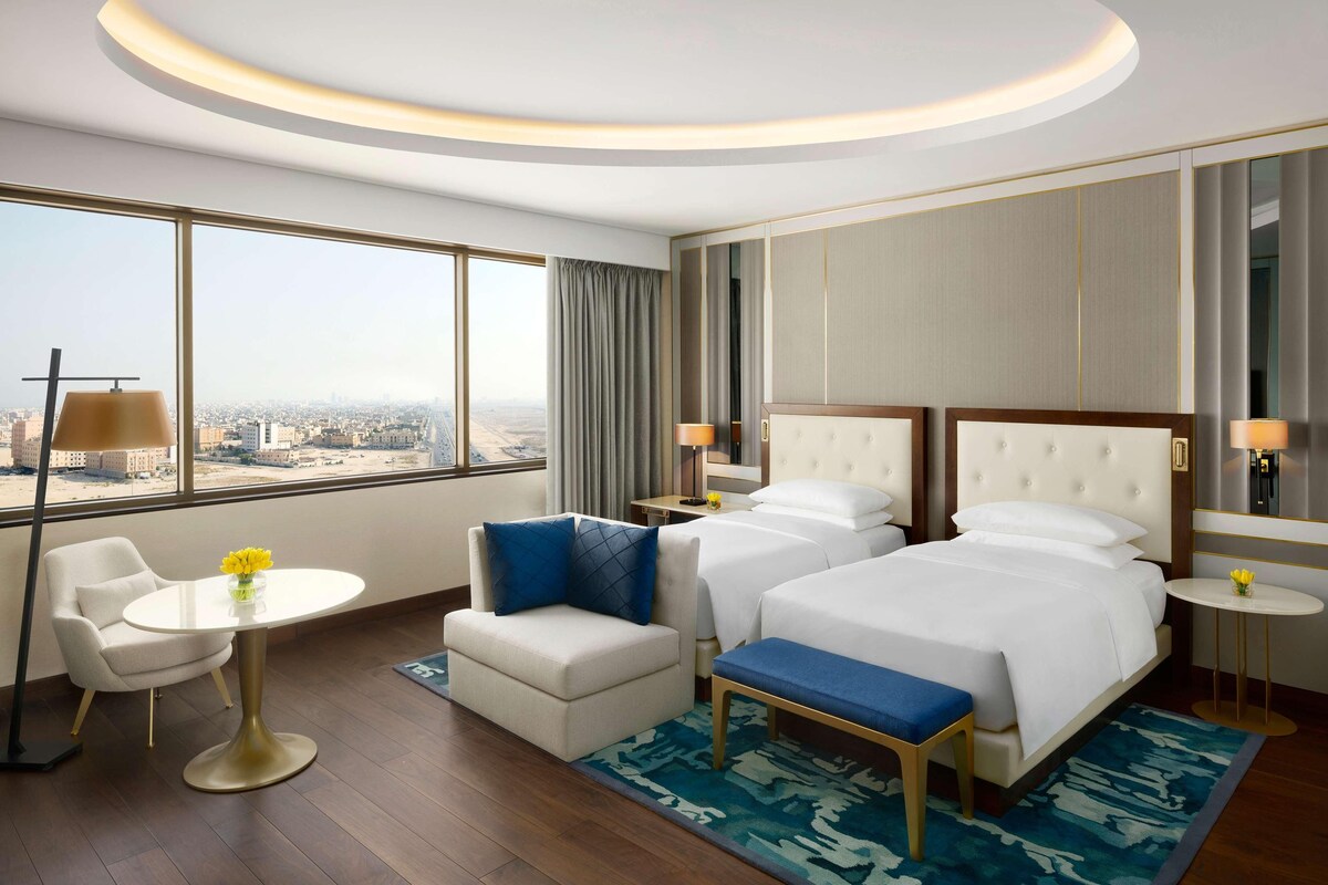 A hotel room with a large window, two beds, a small table, a sofa, and a bench.