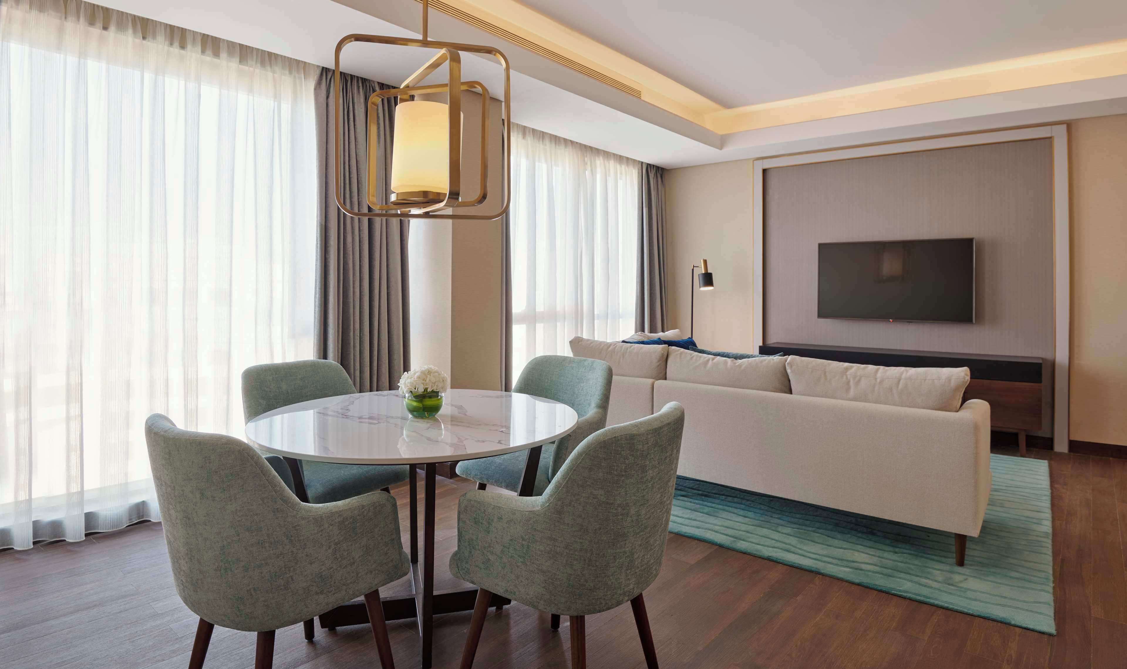 A hotel room with a round dining table, two chairs, a sofa, a TV, and a large window with curtains.