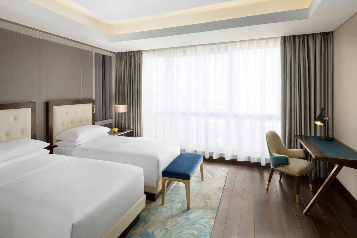 A hotel room with two beds, a desk, a chair, a lamp, and a view of the outside through large windows.