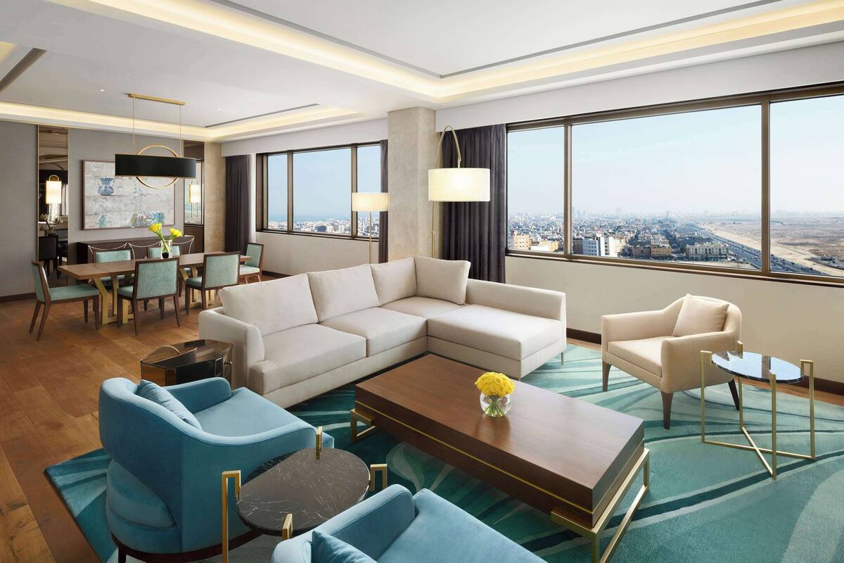 A modern hotel room with a large sectional sofa, a blue armchair, a coffee table, and a dining area with a table and chairs.