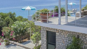 En bygning i sten med en terrasse med udsigt over vand, en pergola og udendørs møbler med parasoller.