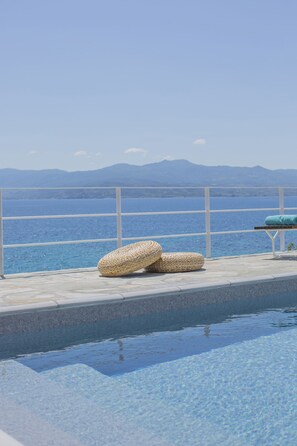 Zona junto a la piscina con vistas al mar y a las montañas. Hay dos esteras tejidas y un banco con cojín.