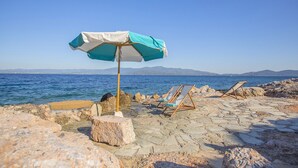 En strand med en stenbelagt plads, en blå og hvid parasol og træloungestole.