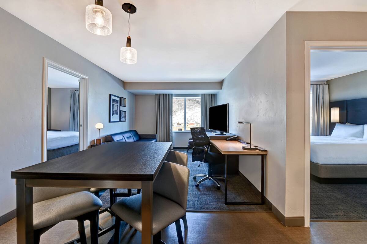 A hotel room with a large bed, a desk with a computer, a dining table with chairs, and a TV.