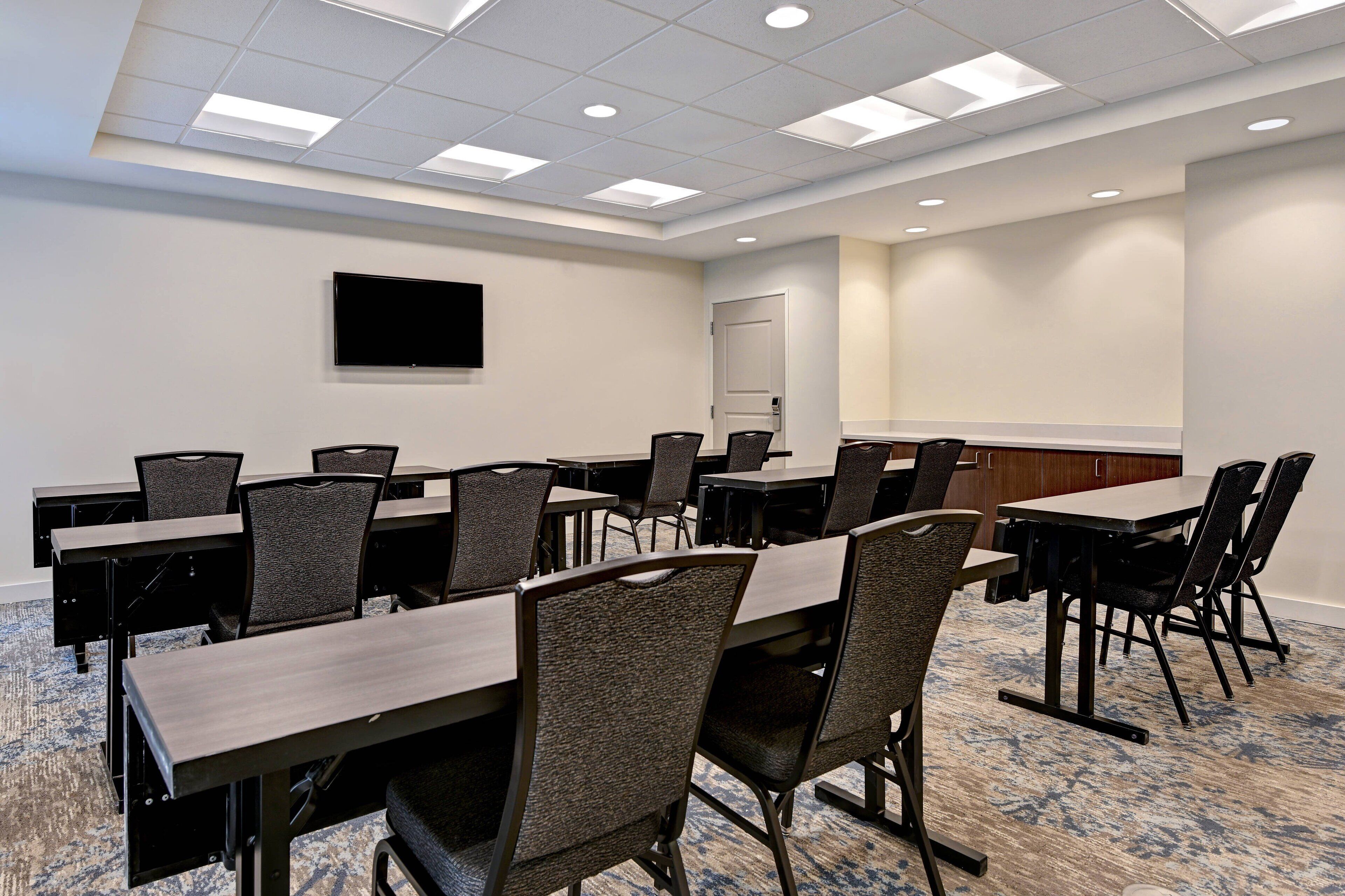 Une salle de conférence avec plusieurs tables et chaises, un téléviseur à écran plat fixé au mur et une porte à l’arrière-plan.