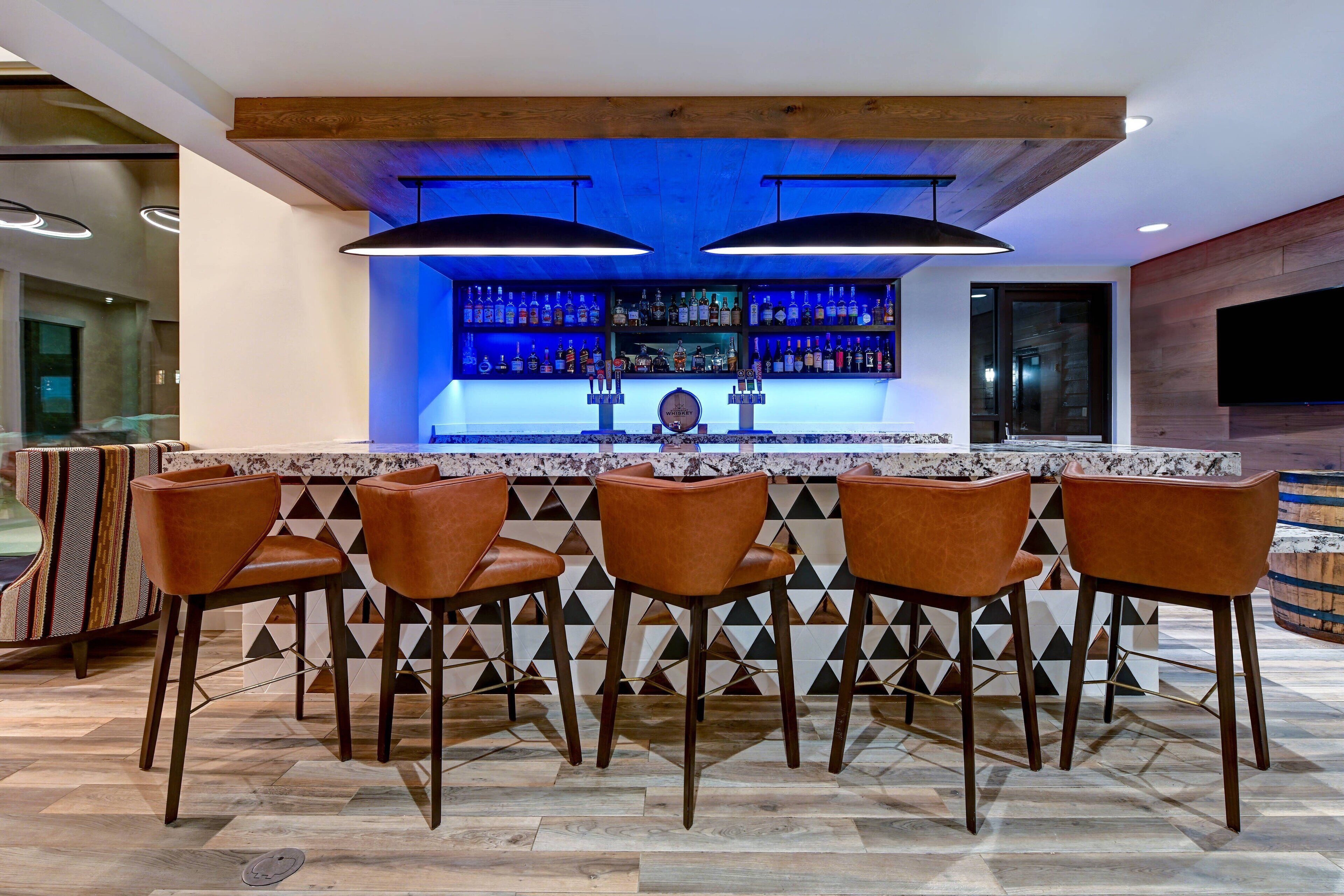 Zona de bar con encimera de mármol, sillas de madera y estante iluminado en azul con botellas.