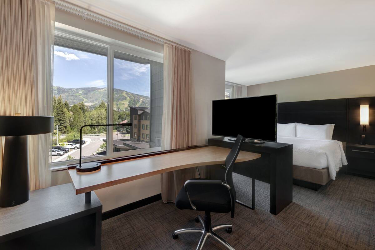 A hotel room with a large window, a desk with a computer, a bed, and a TV.