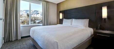Una habitación de hotel con una cama grande, una mesita de noche, una lámpara y vistas a montañas por la ventana.
