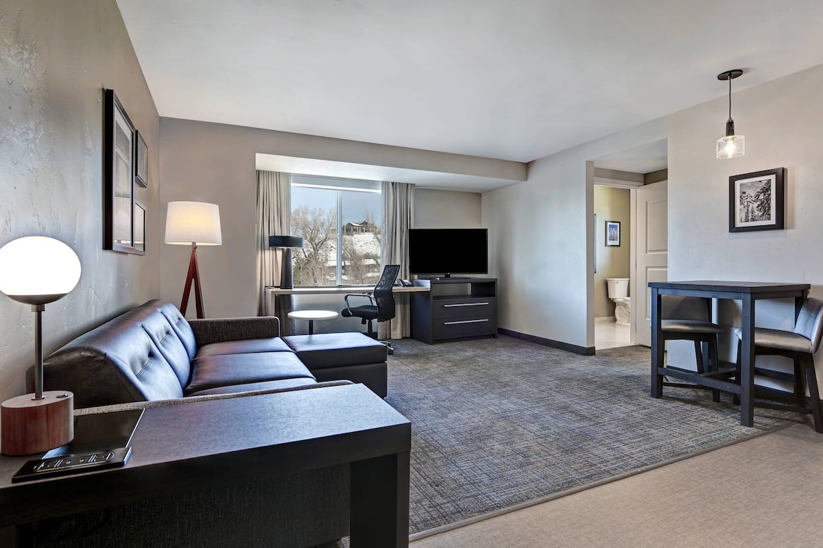A modern hotel room with a sofa, a desk, a flat-screen TV, and a dining area.